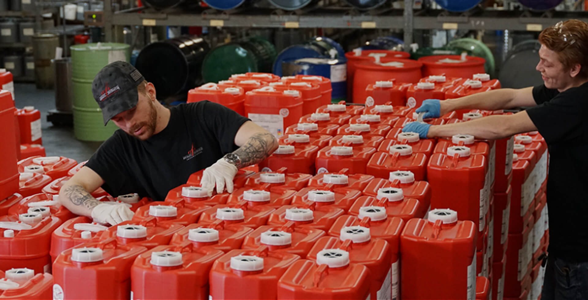 Two AFI warehouse workers wearing gloves labeling and sealing large red fragrance oil containers on a pallet for bulk order fulfillment
